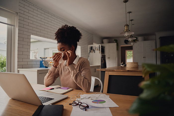 woman blowing her nose while working from home. Her desk is littered with used tissues and it looks like she's suffering from seasonal allergies.